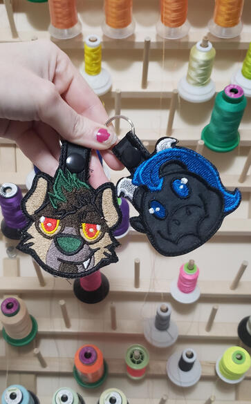 Untitled two head keychains, a hyena with green hair and yellow eyes on the left, and a dragon with blue hair and blue eyes on the right against a background of thread spools.